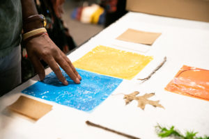 Various objects arranged on a white table, several colored films, a leaf, a small twig- a dark-skinned hand touches a blue square