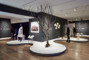 In the galleries of Cooper Hewitt, a large bare tree is mounted in a white platform. In the background, a mannequin wearing a white dress is staged next to a digitally rendered tree in bloom with pink flowers. A visitor stands near a crystalline stool made of sea salt.