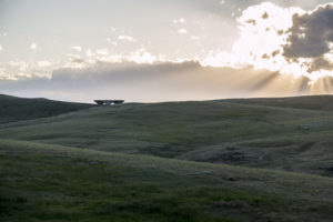The sun shines through clouds upon verdant green hills. In the distance, a large stone sculpture can be seen.