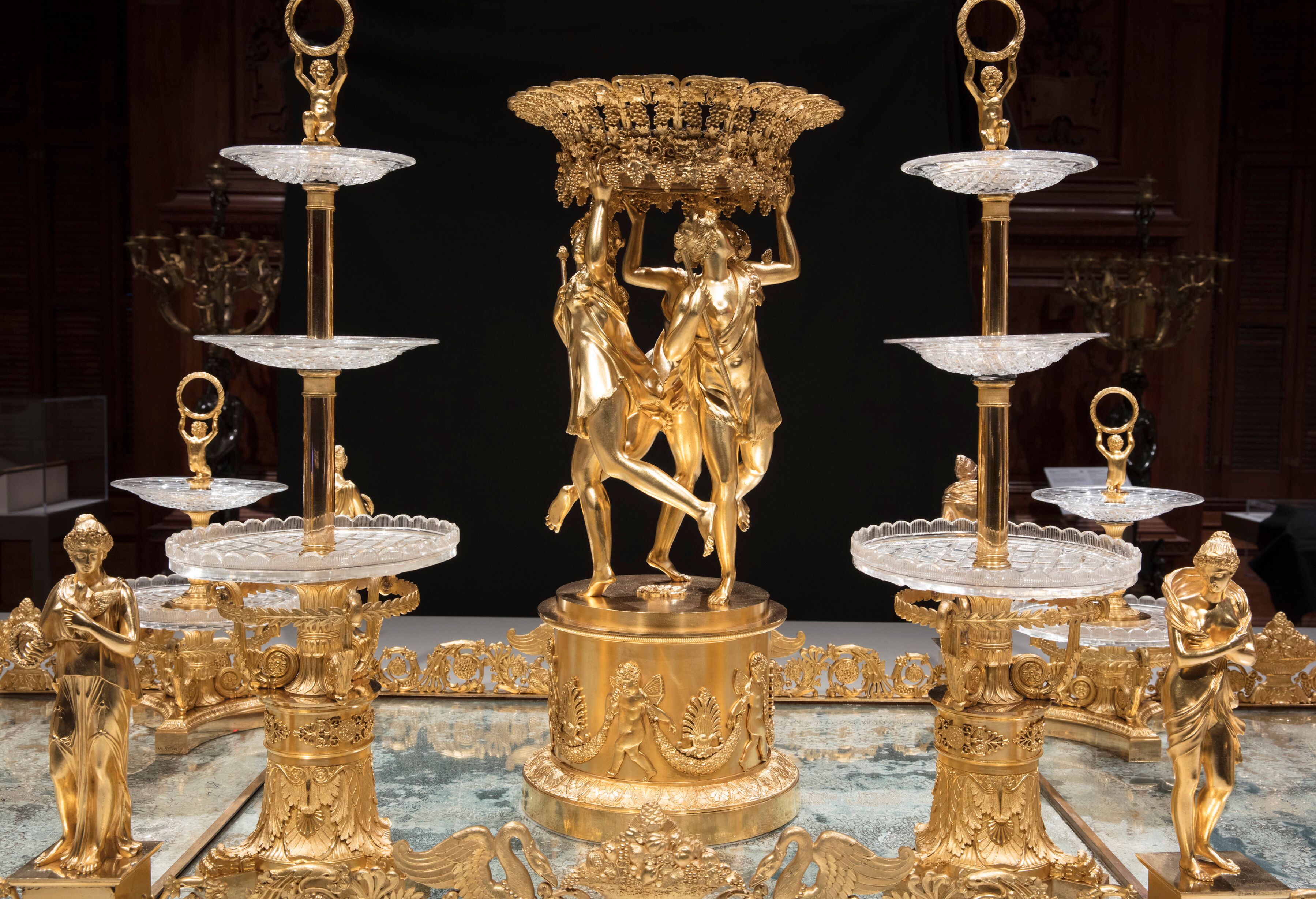 A horizontal photograph of an elaborate gold and glass table centerpiece, against a dark background, focusing on a central sculpture of three women and two towers of glass dishes.