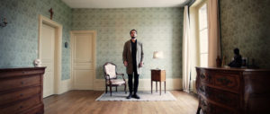 Man stands across from us in a wallpapered room atop an area rug between a chair and end-table