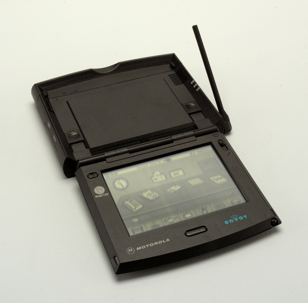 Image features square black form, hinged at the top and opened to reveal an LDC screen showing a black and white drawing of an office with clock and in and out boxes on the wall, a file cabinet, and a desktop in the foreground having a telephone, rolodex, letter, pad, and calendar. A row of icons for different functions is below the desk. A short antenna is attached on right side of the lid, near the hinge. Please scroll down to read the blog post about this object.