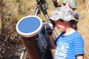 A white five year old boy wearing a camouflage bucket hat peers through a sun scope, a telescope like device. It is in the middle of the day and the sun is shining brightly.