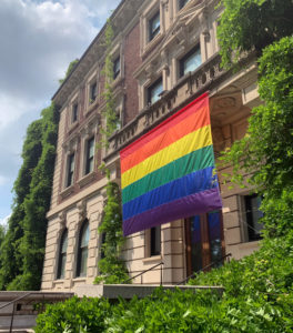 South-facing edifice of beautiful Carnegie Mansion with a rainbow pride flag draped from a second floor balcony on bright summer day