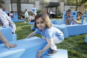 In Cooper Hewitt's Arthur Ross Terrace and Garden, a smiling three year old girl with big dark eyes and short brown hair tries grasps a blue foam block that's larger than she is. Behind her, more children, about four and five years of age, are building constructions of their own with the blocks