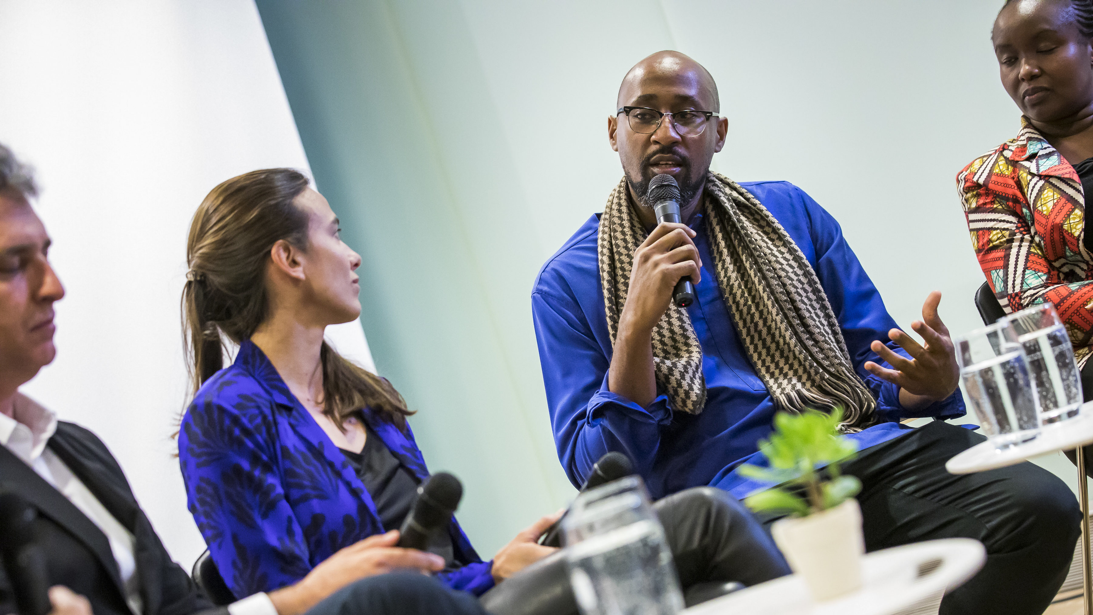 Image of panelists on the stage at Cooper Hewitt, from Ensamble Studios and from Cave architects. The people on the left are in a black suit and vibrant blue woman's suit coat. The two no the right wear colorful African/Kenyan dress, in royal blue and orange patterned.