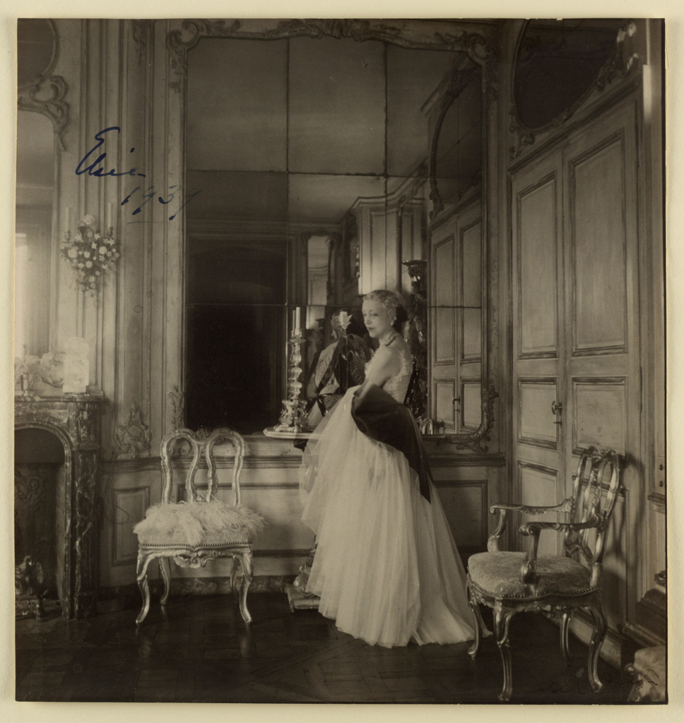 Image features a sepia-tinted photograph of a woman in a long gown standing before a mirrored wall in an elaborate room, a gilded and upholstered side chair to her left and armchair to her right. Please scroll down to read the blog post about this object.