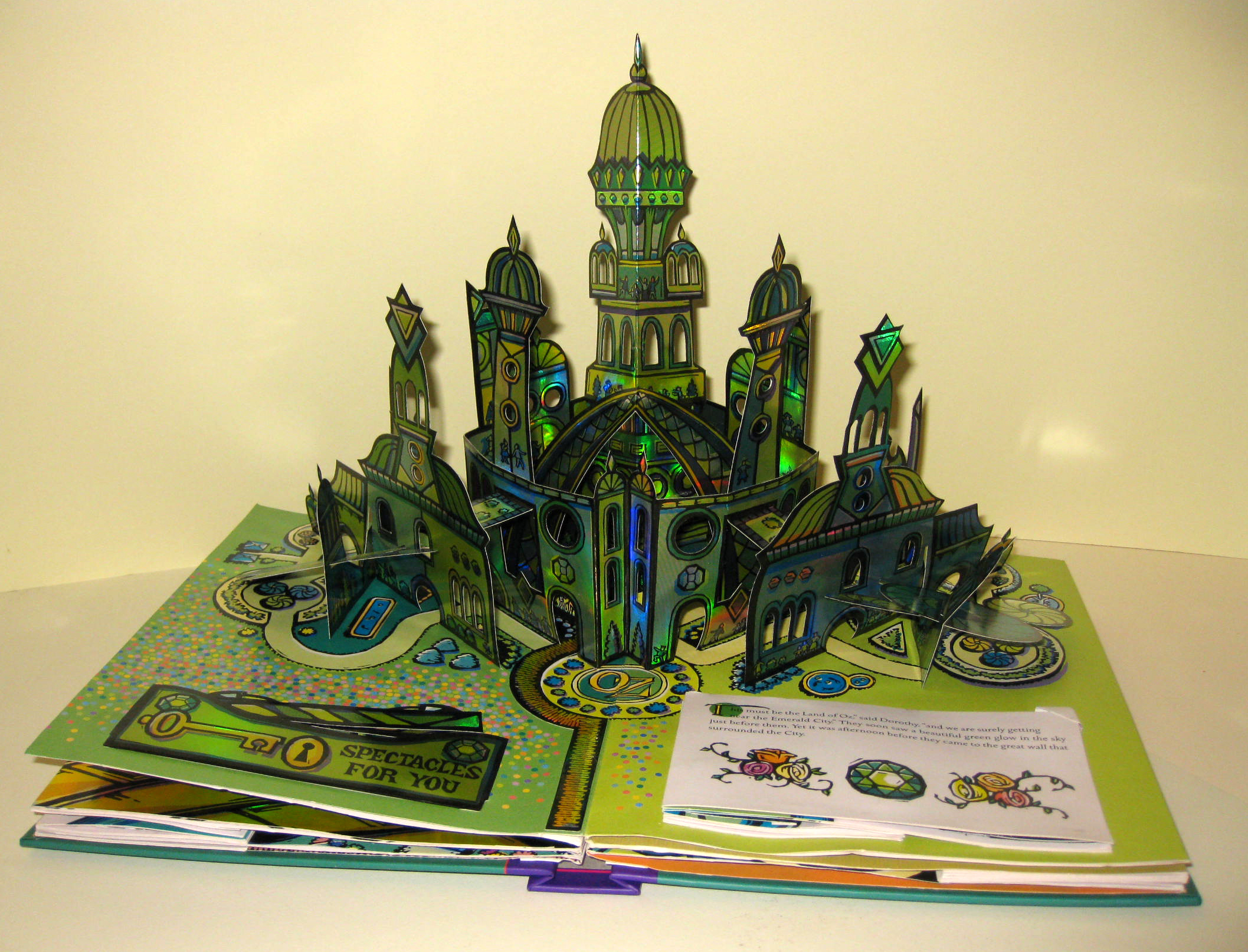 Photograph of a pop-up book. The book is open to to green spread, from which arises a number of green buildings and towers to construct the Emerald City from the Wonderful Wizard of Oz.