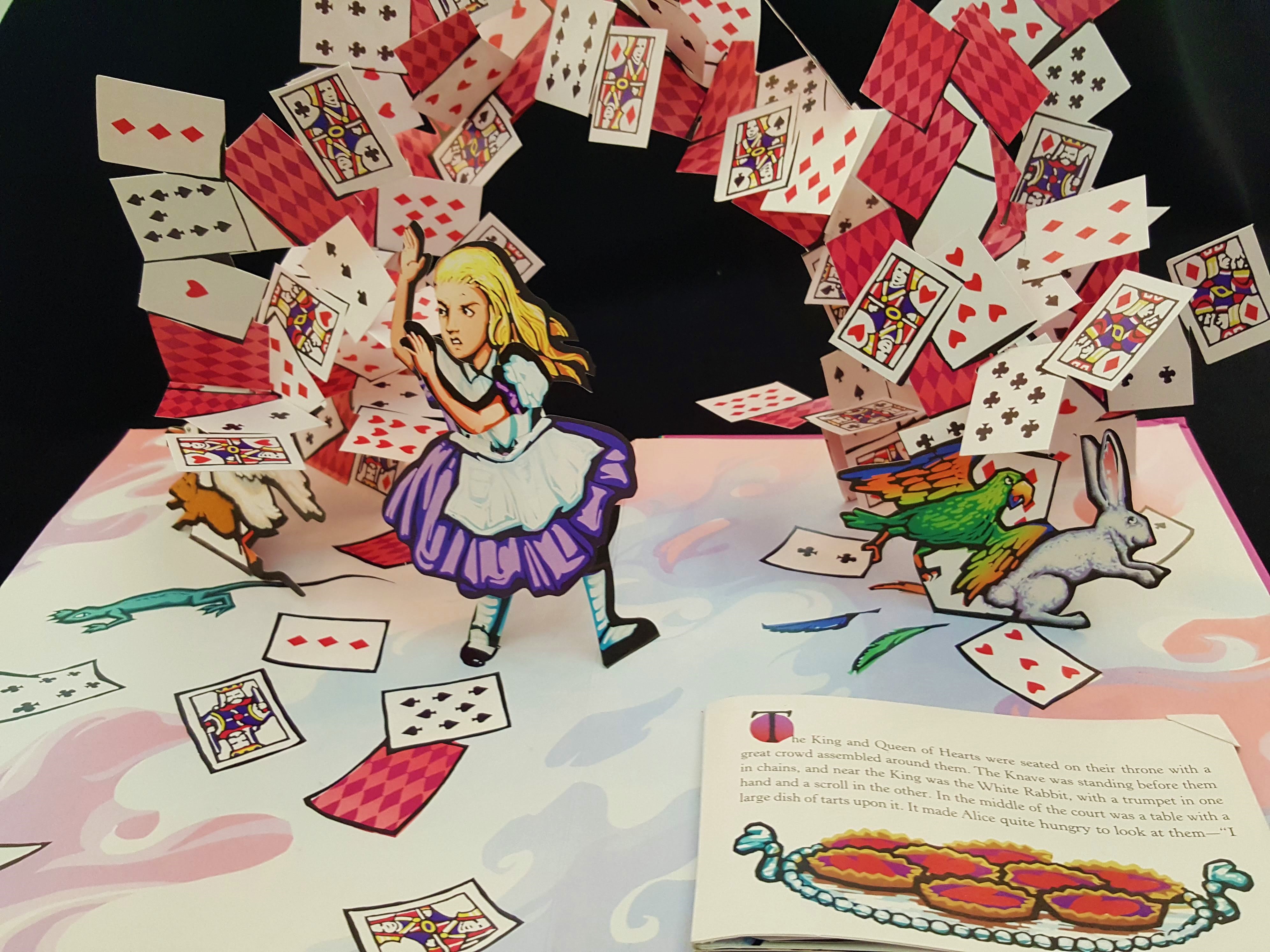 Image shows a page spread from the pop-up book, Alice's Adventures in Wonderland. On a colorful ground, an arch of playing cards looms over a small girl with blonde hair in a purple-blue dress. Please scroll down to read the blog post about this object.