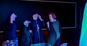 Matilda McQuaid explains to three visitors wearing paper glasses, similar to 3D glasses, how the dress they are looking at appears to glow blue