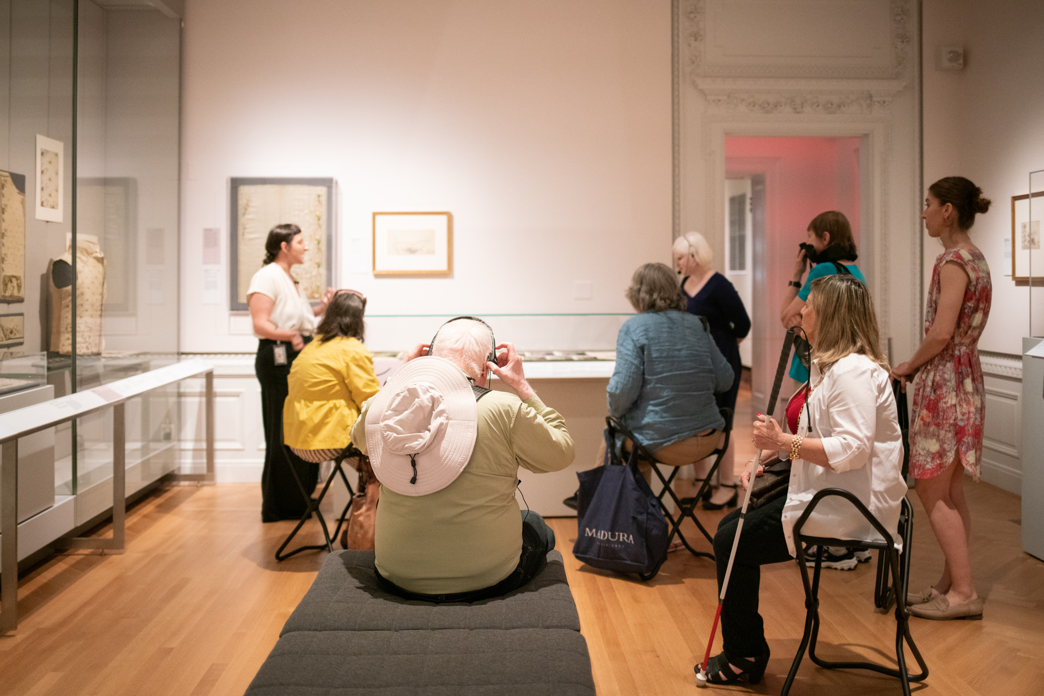 Eight people in a brightly lit gallery at Cooper Hewitt, seated in front of a glass case during a monthly visual description tou