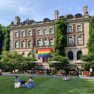 On a beautiful summer day, a rainbow flag hangs from the second floor balcony on the south-facing edifice of lovely Carnegie Mansion. Museum-goers gather on the verdant green lawn