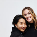 An Asian women with short black hair and a white woman with curled blonde hair stand together smiling at the camera.