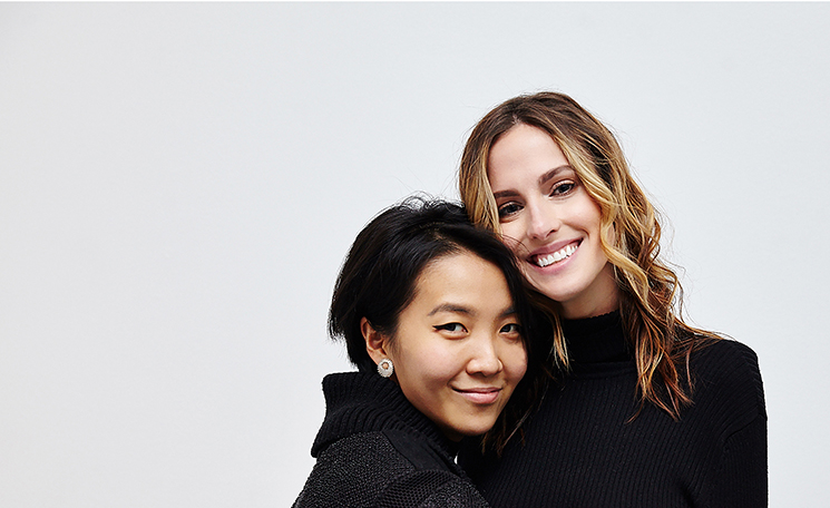 An Asian women with short black hair and a white woman with curled blonde hair stand together smiling at the camera.