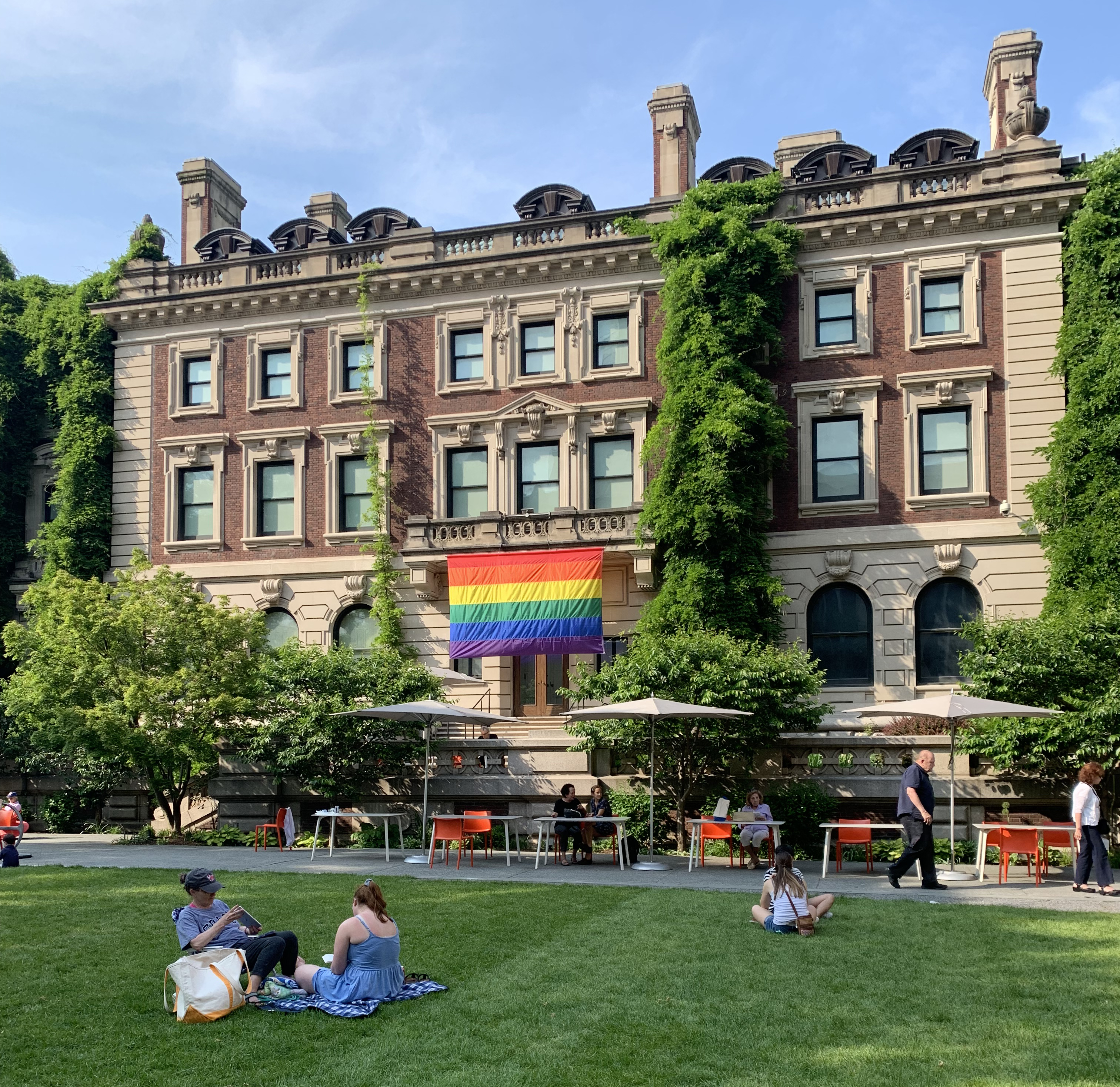 A photograph of a large brick and stone building that is set against a blue sky and a field of green grass stretched before it. Hung from the middle of the building is a rectangular rainbow flag, composed of six stripes of red, orange, yellow, green, blue, and purple. People congregate in small groups on the grass.