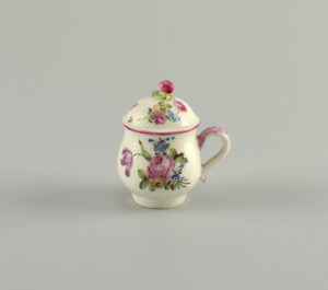 Image features a full-bellied cup with spiral ribbing, a simple loop handle, and a domed lid with a knob at the top in the form of an apple with leaves. The pot and lid are decorated with naturalistic looking flowers in shades of pink, red, blue, yellow and green, all on a white ground. Please scroll down to read the blog post about this object.