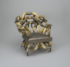 Image features a large armchair, the frame made of multiple curved, twisted, and joined Longhorn steer horns comprising a bow-shaped back and arms surrounding a rectangular seat upholstered in modern metalic-toned leather on curved horn supports and legs terminating in feet with small brass casters. Please scroll down to read the blog post about this object.