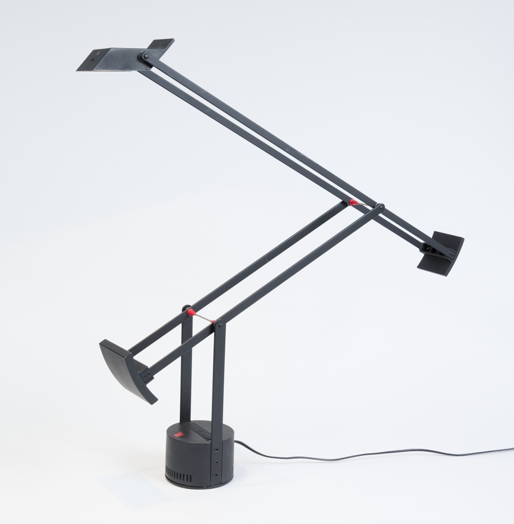 Image features a black desk lamp consisting of a small rectilinear bulb housing with reflector supported by two pairs of counter-weighted adjustable arms set on a swiveling cylindrical base with cooling slots and a red plastic on/off switch. Please scroll down the read the blog post about this object.