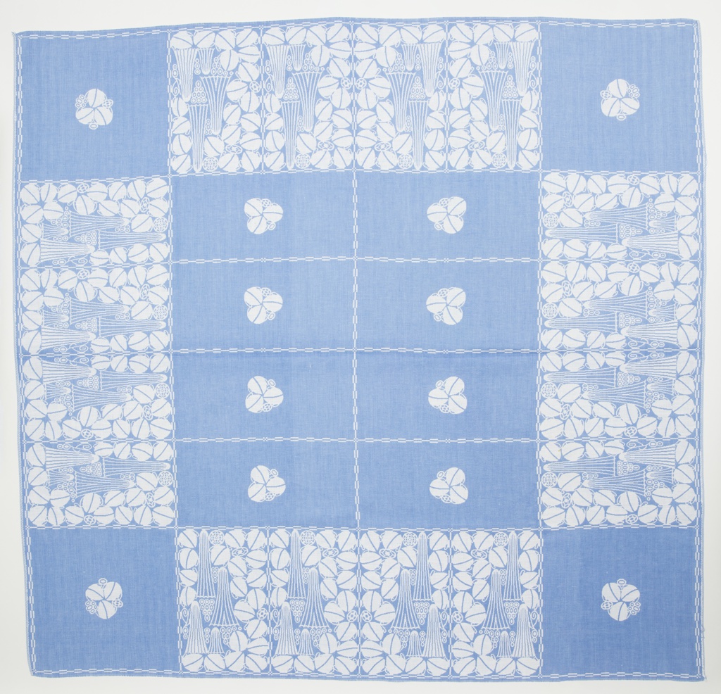 Image features a blue and white tablecloth with a central section containing eight blue rectangles, each with a single stylized floral element in white. Four squares with the same isolated floral motif are at each corner with rectangles between containing a dense arrangement of geometrical and floral elements. Please scroll down to read the blog post about this object.