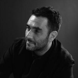 Black and white photograph of Lawrence Azerrad capturing his head down to his upper arm. He glances towards the bottom left corner so that his face is mostly in profile. He bares a small, closed-mouth smile and a 5'oclock shadow.