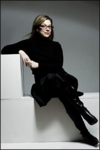 Debbie Millman sits in the middle of an L-shaped white block, with her right elbow resting on the top and legs crossed. She's dressed in all black including her glasses frames. She looks directly at the viewer with an intelligent smirk, hair blowing every so slightly.
