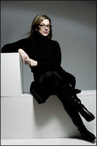 Debbie Millman sits in the middle of an L-shaped white block, with her right elbow resting on the top and legs crossed. She's dressed in all black including her glasses frames. She looks directly at the viewer with an intelligent smirk, hair blowing every so slightly. 