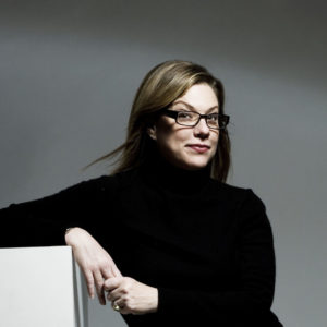 Debbie Millman sits in the middle of an L-shaped white block, with her right elbow resting on the top and legs crossed. She's dressed in all black including her glasses frames. She looks directly at the viewer with an intelligent smirk, hair blowing every so slightly.