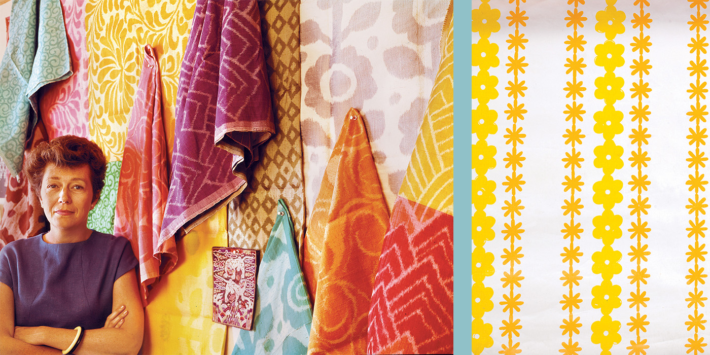 A photograph of designer Gere Kavanaugh leaning against a wall of her vibrantly colored textiles with her arms crossed. She bares a calm expression which parallels the warmth of the wall next to her.