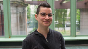 Image of Dr. Max Liboiron looking directly at camera. She has shaved hair on the sides and the top of her hair is pulled back. Her grin is small, but joyful, and her eyes show a smile behind wireframe glasses.