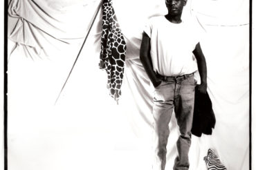A portrait of Willi Smith, hip cocked and looking at the viewer, against a drapey white studio background. He is wearing jeans, a white t-shirt, and sneakers.