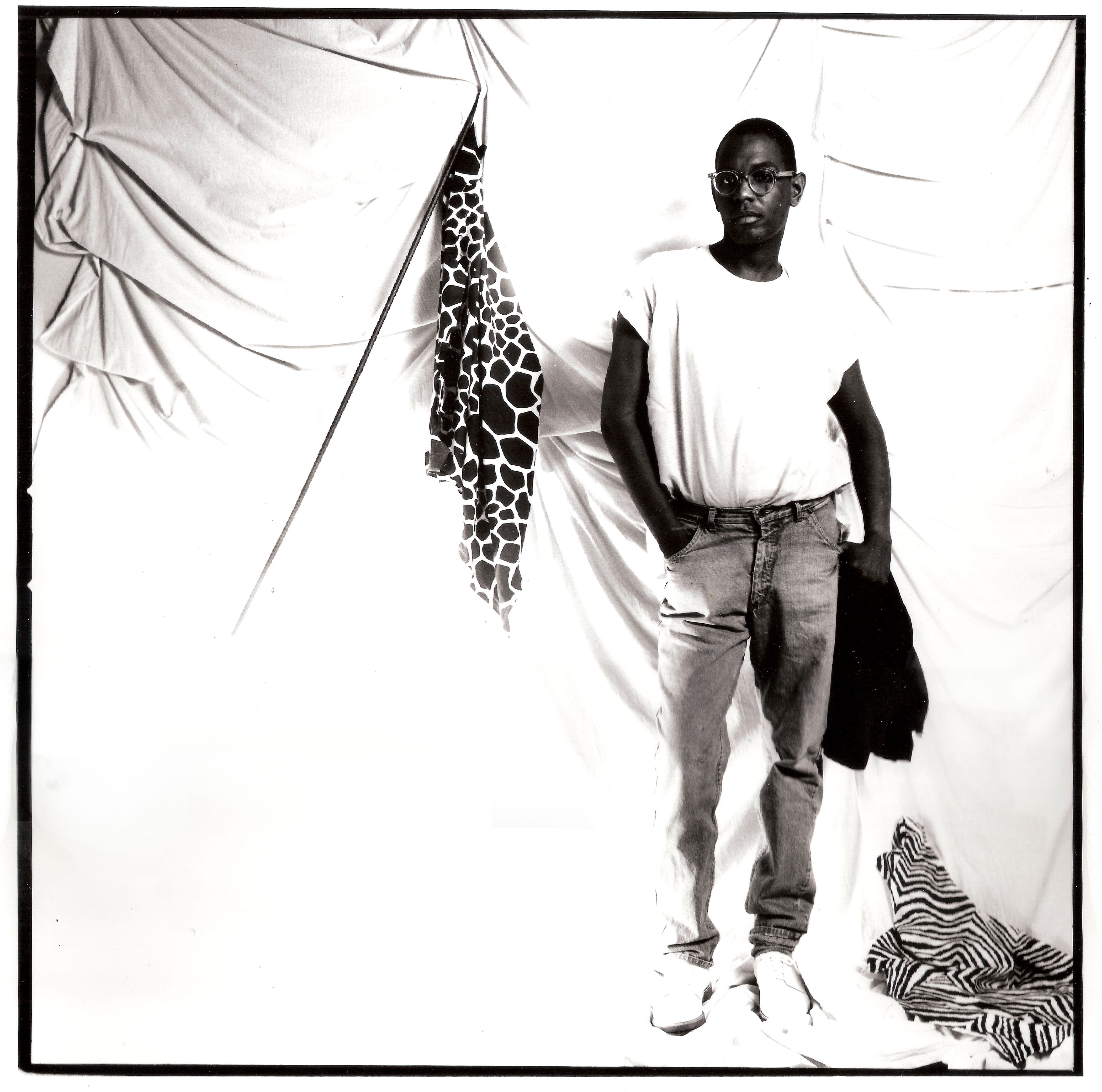 A portrait of Willi Smith, hip cocked and looking at the viewer, against a drapey white studio background. He is wearing jeans, a white t-shirt, and sneakers.