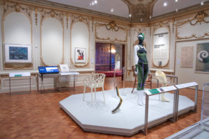 In Cooper Hewitt's gallery, a mannequin wearing a stretchy green athletic suit (including a ski mask) is on display. The mannequin is presented alongside a white chair with branch-like form, a minimalist grey and yellow prosthetic leg, and a white prosthetic leg that is bulky. The gallery is white with historic, ornate gold decorations on the wall and ceiling.