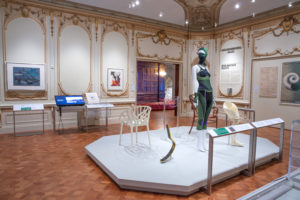 In Cooper Hewitt's gallery, a mannequin wearing a stretchy green athletic suit (including a ski mask) is on display. The mannequin is presented alongside a white chair with branch-like form, a minimalist grey and yellow prosthetic leg, and a white prosthetic leg that is bulky. The gallery is white with historic, ornate gold decorations on the wall and ceiling.