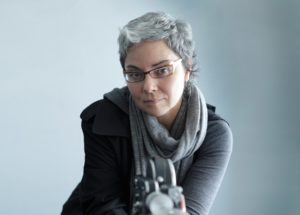 Rebeca Méndez, a woman with light tan skin, short, swooping grey hair, and striking gold glasses, looks directly at the camera with a piercing gaze.