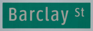 A green, New York City street sign composed of a landscape-orientation rectangle with "Barclay St" in white letters. "Barclay" is largest, while "St," next to it appears smaller, like an exponent. The material of the sign will reflect light, and appears in this image with a small diamond pattern, like a chain-link fence.