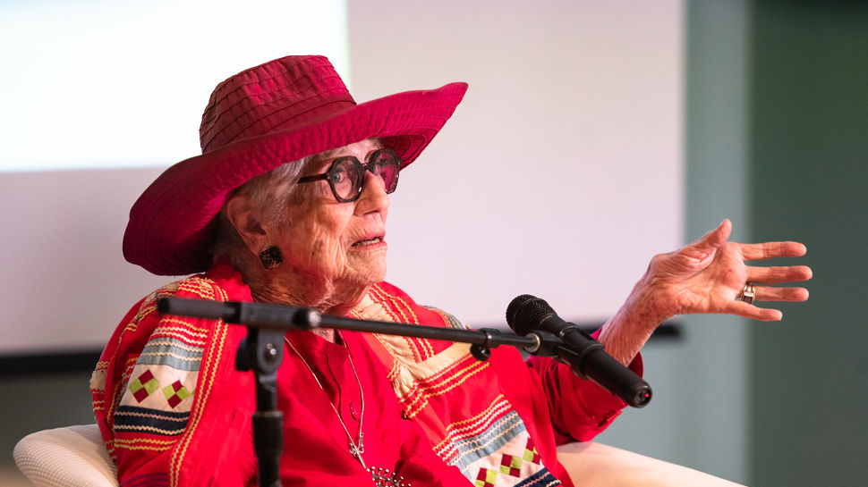 image of Gere Kavanaugh wearing a read sun hat and red dress. She sits with a microphone on a stand in front of her. Her left hand is outstretched and she wears dark rimed glasses