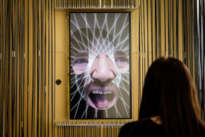 A woman interacts with a screen where blobby cut outs of facial features float around with white lines going everywhere