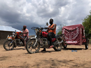On a dirt road, two dark-skinned African men sit atop motorcycles. Hitched to the back of one man's motorcycle is a fabric-covered tent on wheels. The fabric is red and is printed with the word "Ambulance."