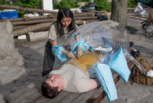 This is a photo demonstrating a surgical device for poverty areas. A man lies on his back with his eyes clothes. His torso is covered with an inflated balloon. A young woman pantomimes surgery on the man. Her hands are in the bubble (they pass through small side openings) but her arms and body are outside the bubble.