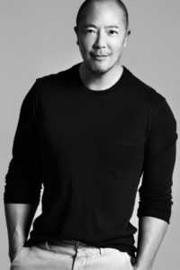 Black and white photo of Derek Lam. He's a thirty or forty-something year old Asian-American man who has a shaved head. He's wearing a long sleeved black tee shirt and is slightly smiling.