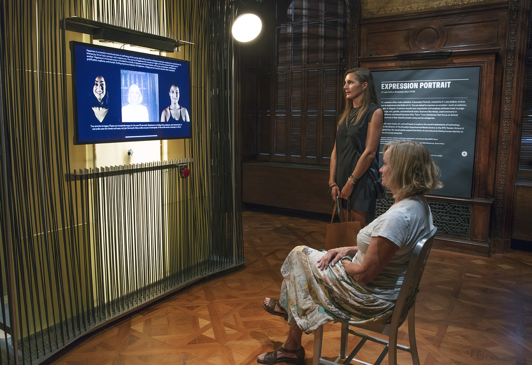 Two women, one seated and one standing, are positioned in a gallery space. They both look intently at a video monitor affixed to a wall covered in dozeons of black vertical rods. The monitor shows text, images of two women, and a live feed of the seated onces facial expressions.