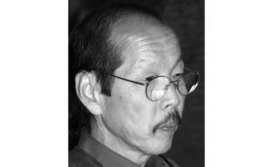 A black and white photograph of Frank Ching in profile. He has a mustache and round glasses.