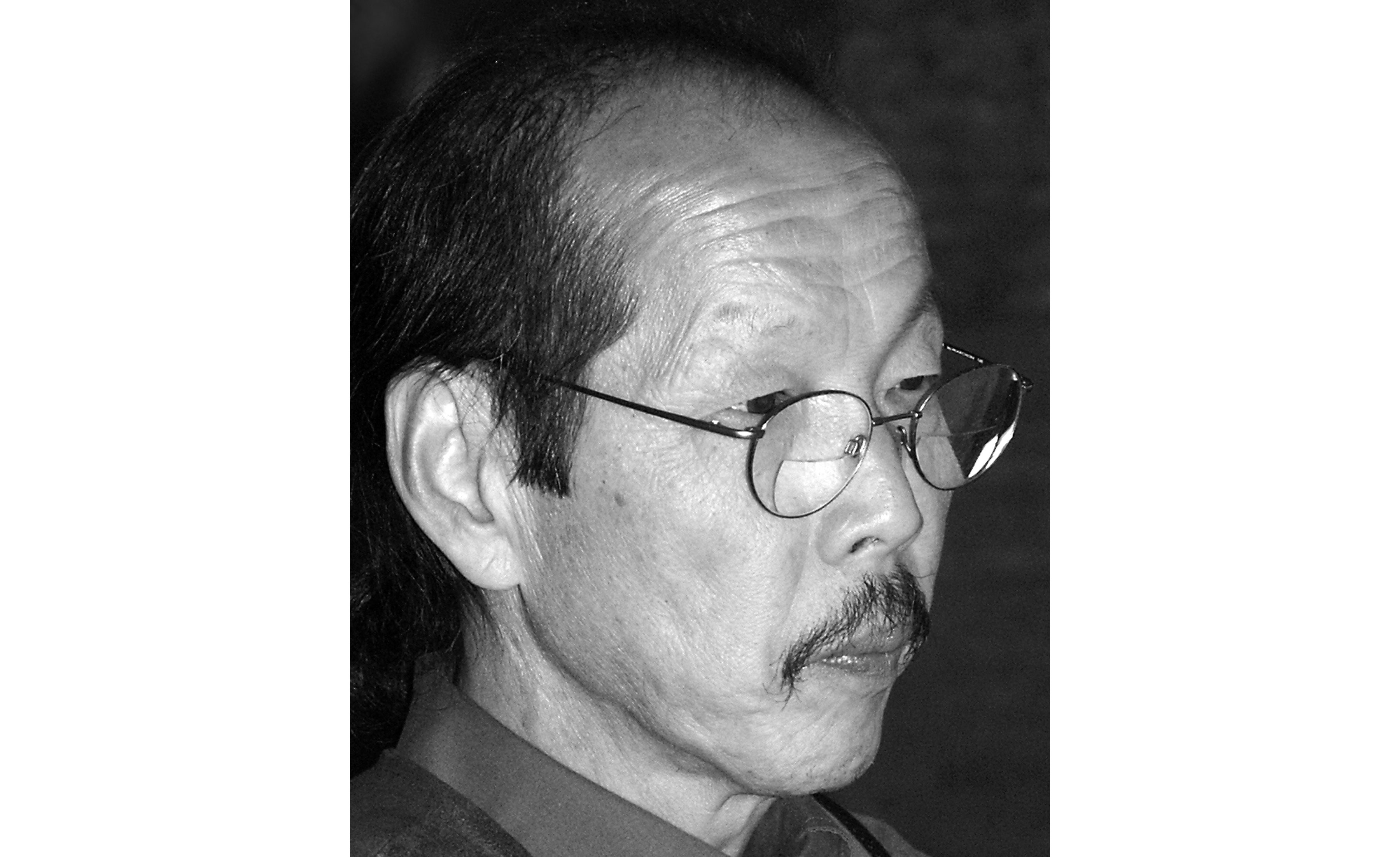 A black and white photograph of Frank Ching in profile. He has a mustache and round glasses.