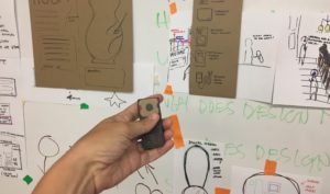 A hand holds a small cardboard prototype in front of a wall covered in drawings and notes