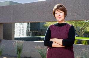 Patricia Moore, a white woman with a brown bob haircut in her sixties, stands near an architectural waterfall. Her arms are folded and her head tilted forward. She's wearing a purple tunic layered over a 3/4 sleeve length black tee.