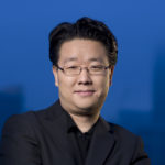 A photograph of Jeff Han in a black shirt with a black suit jacket and thin rimmed glasses. Behind him is a blue background.