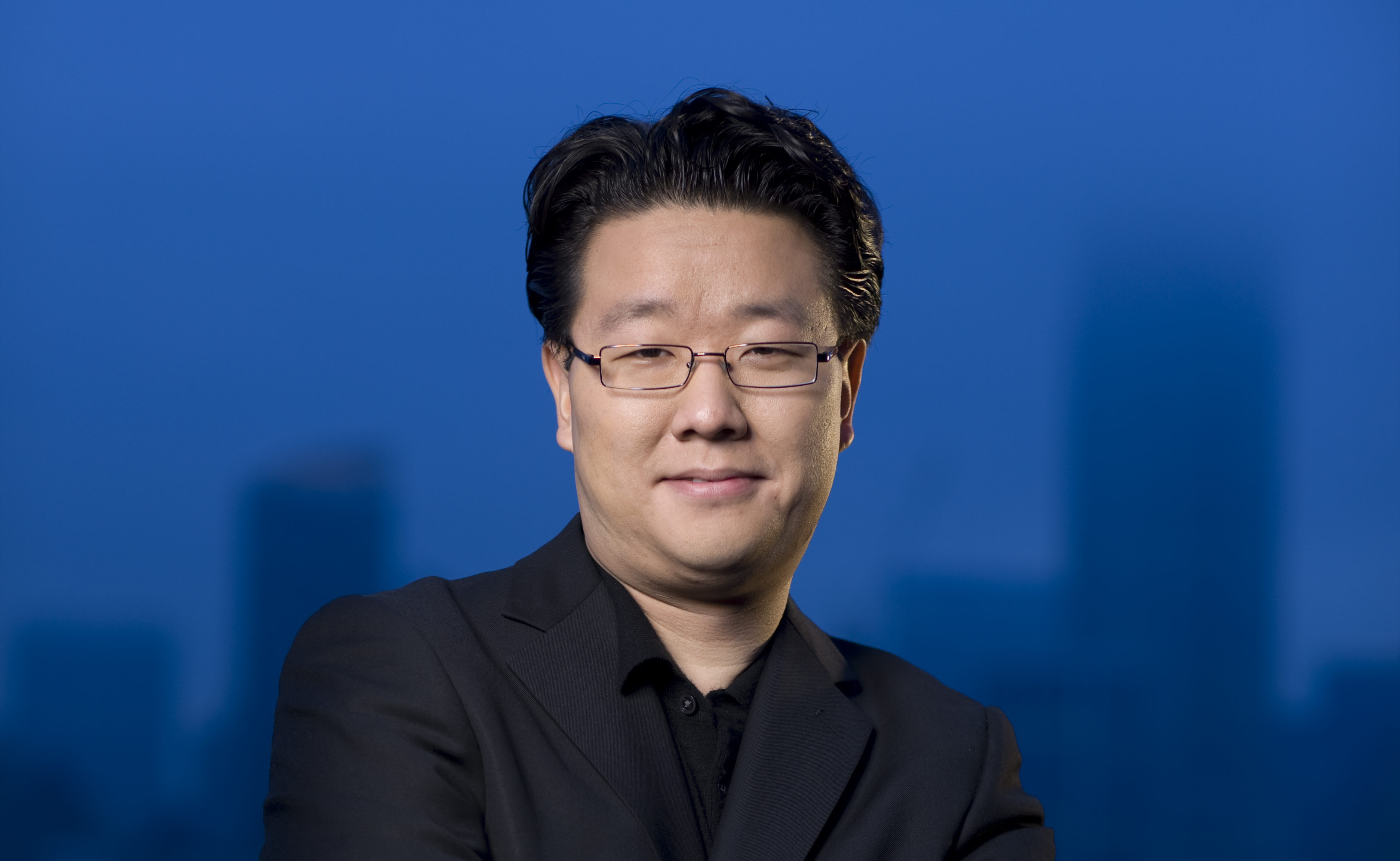 A photograph of Jeff Han in a black shirt with a black suit jacket and thin rimmed glasses. Behind him is a blue background.