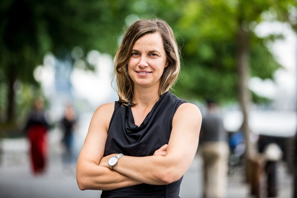 Kate stands with her arms folded in an outdoor public space. She is blonde, tan, in her thirties, and has strong arms. She is wearing a black tank top and silver watch. She is smiling wryly.