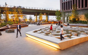 In an urban plaza, children play on artificial "stepping stones" on a water feature made from concrete and ringed by golden light. A man in a suit walks by. Another man lounges on a wooden bench and looks at his phone. There is an elevated highway in the near background.