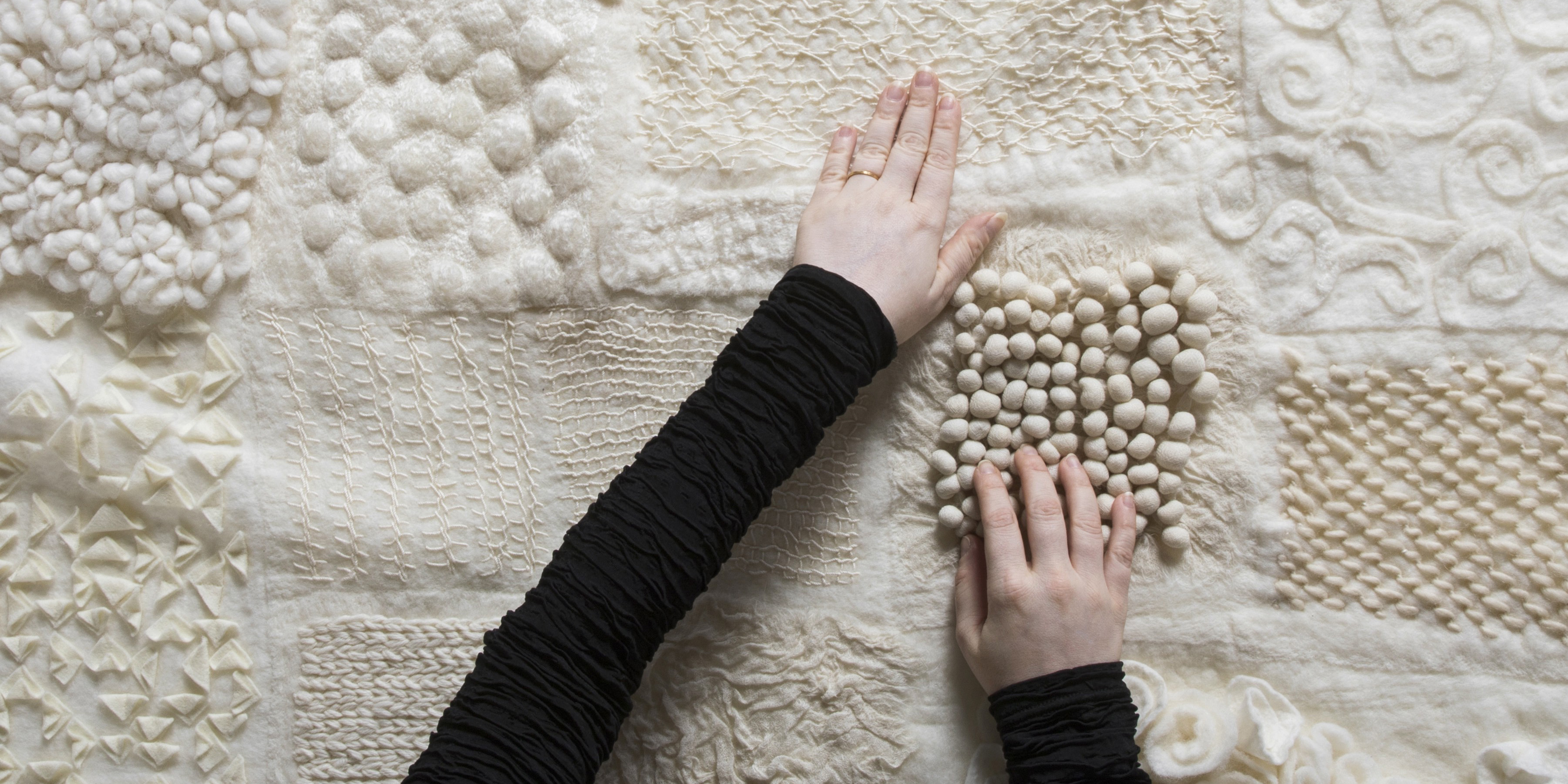 Textile Workshops Drop in on Design, Photograph from above of a patchwork textile made of felt and embroidery techniques, in shades of white. Two hands feel the cloth.